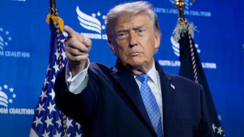 Donald Trump pointing at someone during a press conference, standing against a blue background