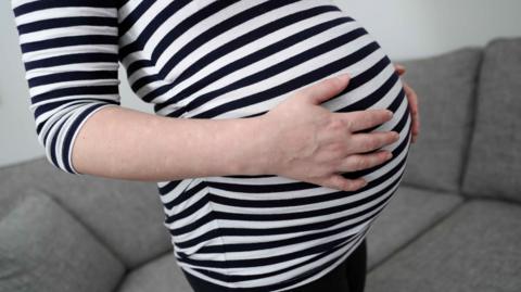 The image shows a close-up view of a pregnant person's bump, with both hands resting on the stomach while wearing a navy and white striped top. In the background there is a grey sofa, and the photograph appears to have been taken indoors using soft natural light, with the focus centred on the pregnancy bump rather than the person's face, which is not visible.