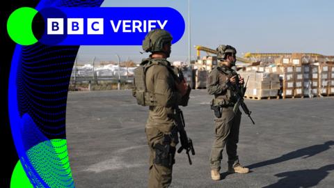 Two Israeli soldeirs stand guard near aid supplies in Gaza. They are wearing tactical gear and carrying rifles.  In the background large pallets with boxes can be seen.