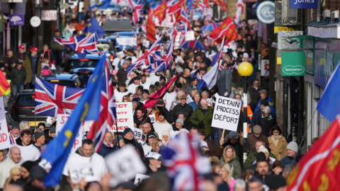 Hundreds of people marching on a town high street. Some people are carrying union jacks and Sussex county flags, while one person holds a sign that reads "detain process deport".
