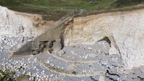An aerial image of brown stone steps leading down a white chalk cliff face. A person in a red top is stood at the top of the steps. Below the cliffs is a pebble beach with large boulders strewn across it. On top of the cliff is green grass.