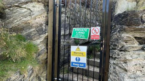Steel gate blocking a narrow cliffside stairway, with signs reading "Private Land", "No entry" and "Keep out" attached to the gate.