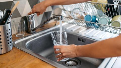A woman holds a glass under a running faucet. It is about halfway filled with water. Dishes can be seen left to dry on a rack to her right.