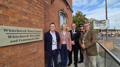 Four men and one woman stand outside a brick building next to a road. Agold sign with black writing on the wall reads "whitchurch town council. whitchurch town hall and community hub". One of the men is a police officer, and is wearing a black police hat and black vest. The woman is the mayor and is wearing a pink top and cardigan with a chain of office around her neck