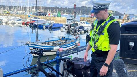 A police officer in a bright yellow vest stands in front of a bike leaned next to a railing. In the background there are boats in a harbour. 