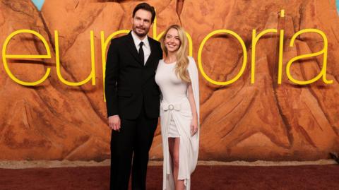 Sydney Sweeney and Sam Levinson standing and smiling together in front of large yellow Euphoria lettering on the red carpet