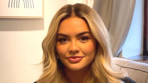 Olivia McVeigh - a young woman with long blonde hair looks at the camera while sat on a grey sofa in a white room