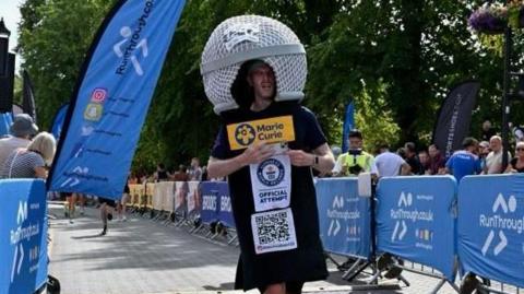 Participant dressed in a large helmet-shaped costume runs along a race course toward the finish area, wearing a Marie Curie charity bib and official attempt sign. Blue event barriers, spectators, flags and a race clock line the route beneath trees on a sunny day.