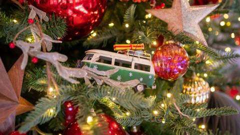 A camper van decoration on a Christmas tree surrounded by red and silver baubles.