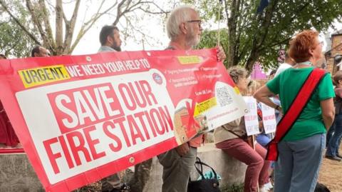 A man holds a red sign that says "URGENT, WE NEED YOUR HELP TO SAVE OUR FIRE STATION". There is a crowd of people behind him. There are also trees in the background. 