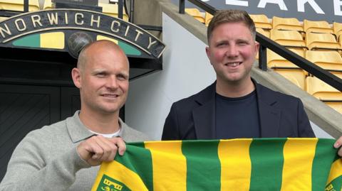 Liam Manning and sporting director Ben Knapper hold their team's scarf in front of a stadium's tunnel with their team's crest above it