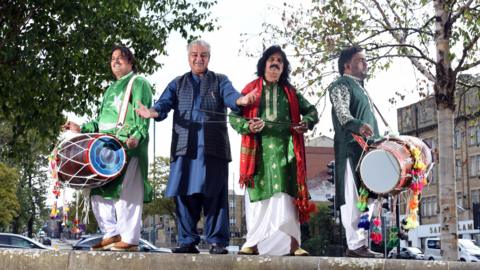 Four South Asian men pictured in an urban streetscape. They are dressed in traditional clothes. Three of them carry dhol drums and other musical instruments.