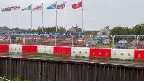 A brown wooden fence and white and red alternating crash barriers run parallel either side of the start-finish straight of a very wet Billown Course. A row of flag poles featuring the Manx, British, Scottish, English, Irish, and Welsh flags flying stiffly in the wind sits behind the barriers. There is also fencing in place in front of cars parked on a grassed area.