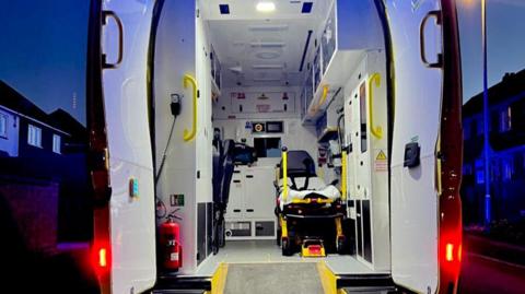 The back of an ambulance with its doors open, showing a bed for a patient inside and cupboards around the edge of the interior. There are houses in the background.