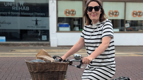 A woman is wearing a white and navy striped dress and shades. She is sitting on a bike with a brown basket on the front. 