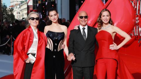 Meryl Streep, Anne Hathaway, Stanley Tucci and Emily Blunt pose on the red carpet in London. Streep is wearing sunglasses, a long, red, satin jacket, a cream blouse and black trousers. Hathaway has her hair up and is wearing a black embellished corset. Tucci is wearing a pinstriped black suit, white shirt and tie, along with sunglasses. Blunt has a red two-piece on an a jewelled necklace.
