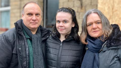 A middle-aged man and woman - both with grey hair - stand in front of a boarded-up house, alongside a young girl who has long, dark hair. They all wear dark-coloured coats and scarves, and there is rain on their jackets.