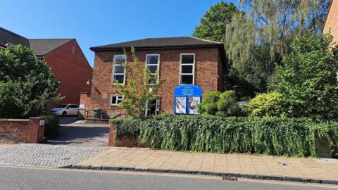 A brown brick building with white windows and a white door. There is a blue sign attached to a brick wall covered in green foliage at the front.