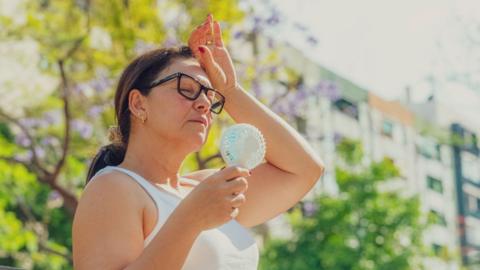 Woman sitting in sun with a battery powered fan, her hand on her forehead