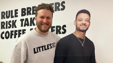 Will Little is wearing a white Little's branded tshirt. He is standing next to a cardboard cut out of Steven Bartlett who is wearing a black top and silver necklace.