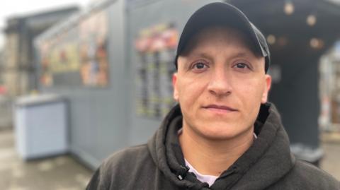 40 year old man wearing a black baseball cap and hoodie in front of a shipping container