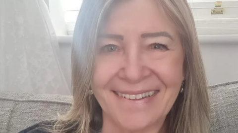 Anne Eason, one of the residents who had to leave the flats, sits on a sofa with a window behind her. She is wearing a cream top and black cardigan and has shoulder-length hair. She is smiling at the camera.
