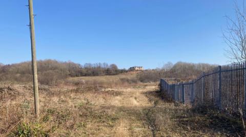 A dry grassed area, with a telegraph pole on the left-hand side, a metal fence on the right and trees in the background. There are houses on a hill in the distance.