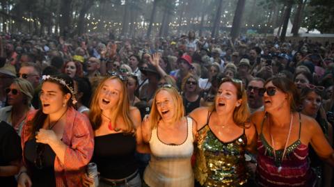 Five women at the front of a packed crowd. They are illuminated by lights from the stage and they have got their arms around each other as they sing along to an artist, who cannot be seen.