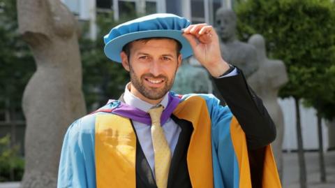 Mark Wood smiles as he looks into the camera. He has dark hair and a thin beard. He is wearing a blue cap and gown.