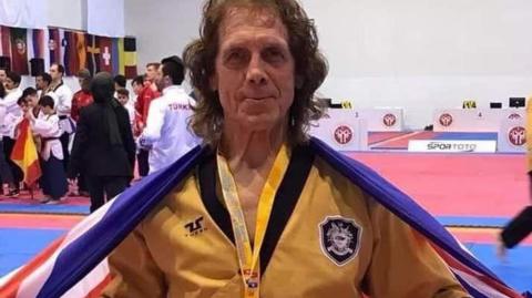 Mike Pejic, an older man with long brown hair and wearing a yellow martial arts outfit, with a medal around his neck, smiles as he stands with a large union flag around his shoulders. Other martial artists can be seen behind him.