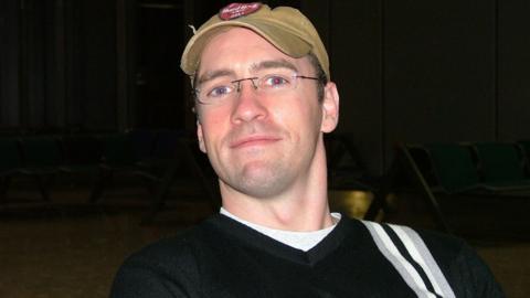 Paul Doyle, then in his late 20s, smiles at the camera while wearing a brown Hard Rock Cafe branded baseball cap and glasses. 