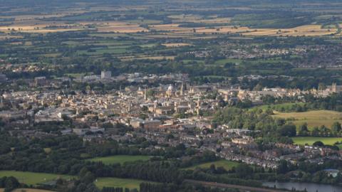 Oxford from the air. The city is surrounded by green fields. Towers and spires can be glimpsed near the centre, with rows of housing at the front and rear.