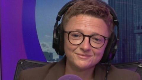 Bev Craig, with short cropped hair and glasses, smiles as she sits behind a microphone wearing headphones