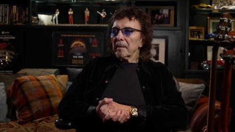 A with dark brown hair, shaded glasses, black jacket and black top, sat on a sofa in a dark room. A guitar is on a stand next to him and there are various ornaments and photos in the background