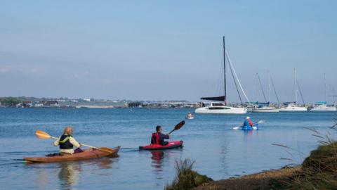 Group of people kayaking on the Christchurch Harbour, Dorset. You can see Mudford spit in the background with a large black building opposite Mudeford Quay. There are three people in kayaks wearing life jackets. There are several yachts moored in the still blue water of the harbour. 