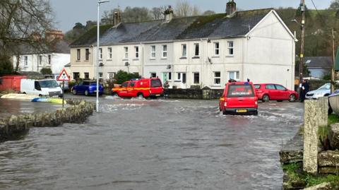 Red vehicles parked at the top of a hill as flood water runs down the road. One of the vehicles, to the right, is moving through the flood water.