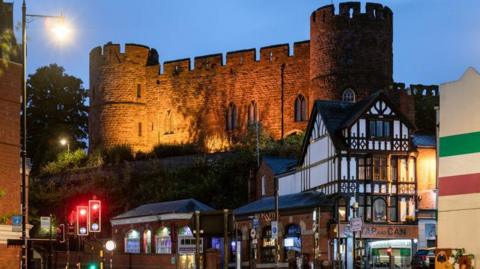 Shrewsbury Castle, a red sandstone building stands on a hill directly above a railway station. There is a black and white Tudor style building in the foreground and traffic lights