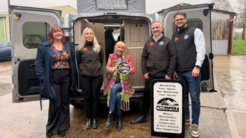 A group of five people stand in front of an open camper van, with a sign for a camper conversion business placed beside them.