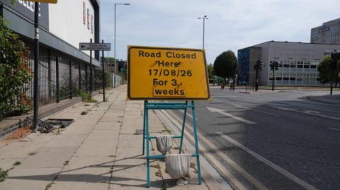 A yellow sign on the pavement says " Road Closed Here 17/08/26 for 3 weeks". The sign is weighted down with sand bags. The road behind it has two lanes with arrows pointing in a direction away from the sign.