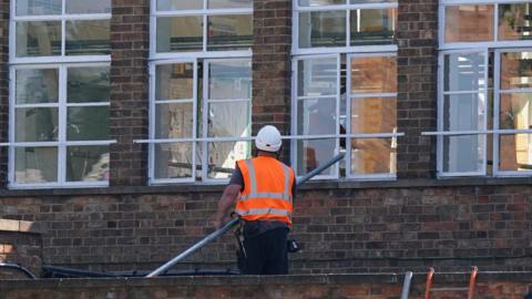 A construction worker in a hard hat and orange safety vest stands outside the brick building of a school, holding a long pole while working on or inspecting the exterior window frames. Several large windows reflect the surroundings, and scaffolding or support bars are positioned across them.