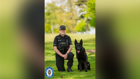 A female police officer in all black uniform kneels beside a black German Shepherd. The pair are pictured in a large open grassy area.