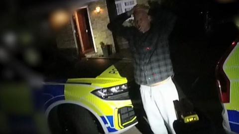 Jeremy Heath has his hands on his head, wearing a dark-coloured check shirt, and standing outside a house in darkness. There are two marked police cars parked either side of him. He appears to have a cigarette in his mouth.