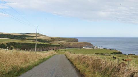 A scenic coastal route where cows are lying down in green fields. There is a house nestled on the coastline in the distance