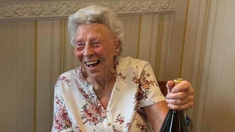 An elderly woman laughs and looks off camera, wearing a white floral blouse