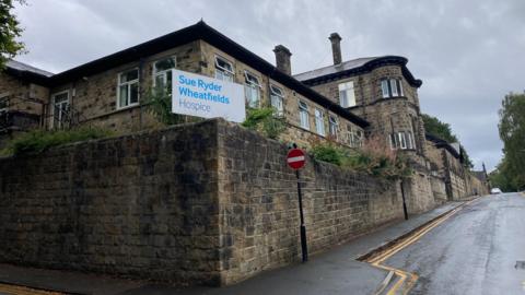 A stone building on the corner of a road with a sign outside which reads 'Sue Ryder Wheatfields Hospice'.