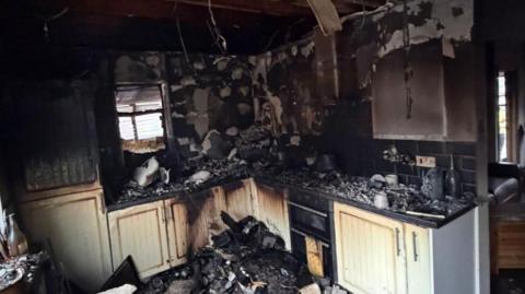 A square-shaped kitchen layout with white cupboards badly burned, charred and damaged by by fire.