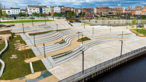 An aerial view of the white steps of the parks amphitheatre which faces on to the river. There are winding paths, slides and spots of greenery dotted around the development. High Street in the background is lined with multi-storey brick buildings in different colours.