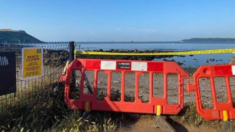 An empty Mount Batten beach near Plymouth which has been closed following the discovery of asbestos. The beach is stoney with lots of brown seaweed on it. The water is dark blue coloured with bright blue skies above it. There is orange fencing and yellow tape blocking access to the beach.