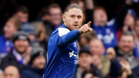 A football player celebrating after a goal. He is pointing his finger out as he runs. He is wearing a blue football shirt, and he has long brown hair that has been tied back.