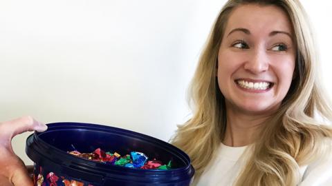 A smiling woman with long blonde hair looking away from a tub of sweets with colourful packaging